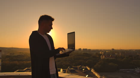 A-male-stockbroker-freelancer-stands-on-a-rooftop-at-sunset-with-a-laptop-and-types-on-a-keyboard-with-his-fingers-looking-at-the-cityscape-from-a-bird's-eye-view.